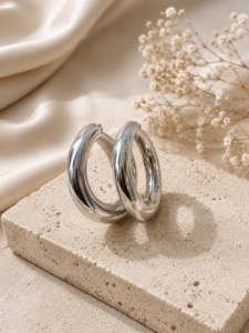 Polished silver chunky hoop earrings displayed on a neutral stone platform with soft beige background.